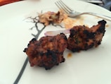 Two pieces of grilled meat are placed on a white plate, showing a slight char on the exterior. A fork rests nearby on the plate, which has a simple black design. There is a hint of garnish, possibly onion, next to the meat.
