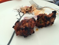 Two pieces of grilled meat are placed on a white plate, showing a slight char on the exterior. A fork rests nearby on the plate, which has a simple black design. There is a hint of garnish, possibly onion, next to the meat.