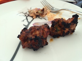 Two pieces of grilled meat are placed on a white plate, showing a slight char on the exterior. A fork rests nearby on the plate, which has a simple black design. There is a hint of garnish, possibly onion, next to the meat.