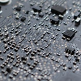 A close-up view of an electronic circuit board filled with numerous small components including microchips, resistors, and capacitors. The surface is covered in a grid-like pattern with intricate metallic traces and soldered connections.