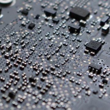 A close-up view of an electronic circuit board filled with numerous small components including microchips, resistors, and capacitors. The surface is covered in a grid-like pattern with intricate metallic traces and soldered connections.