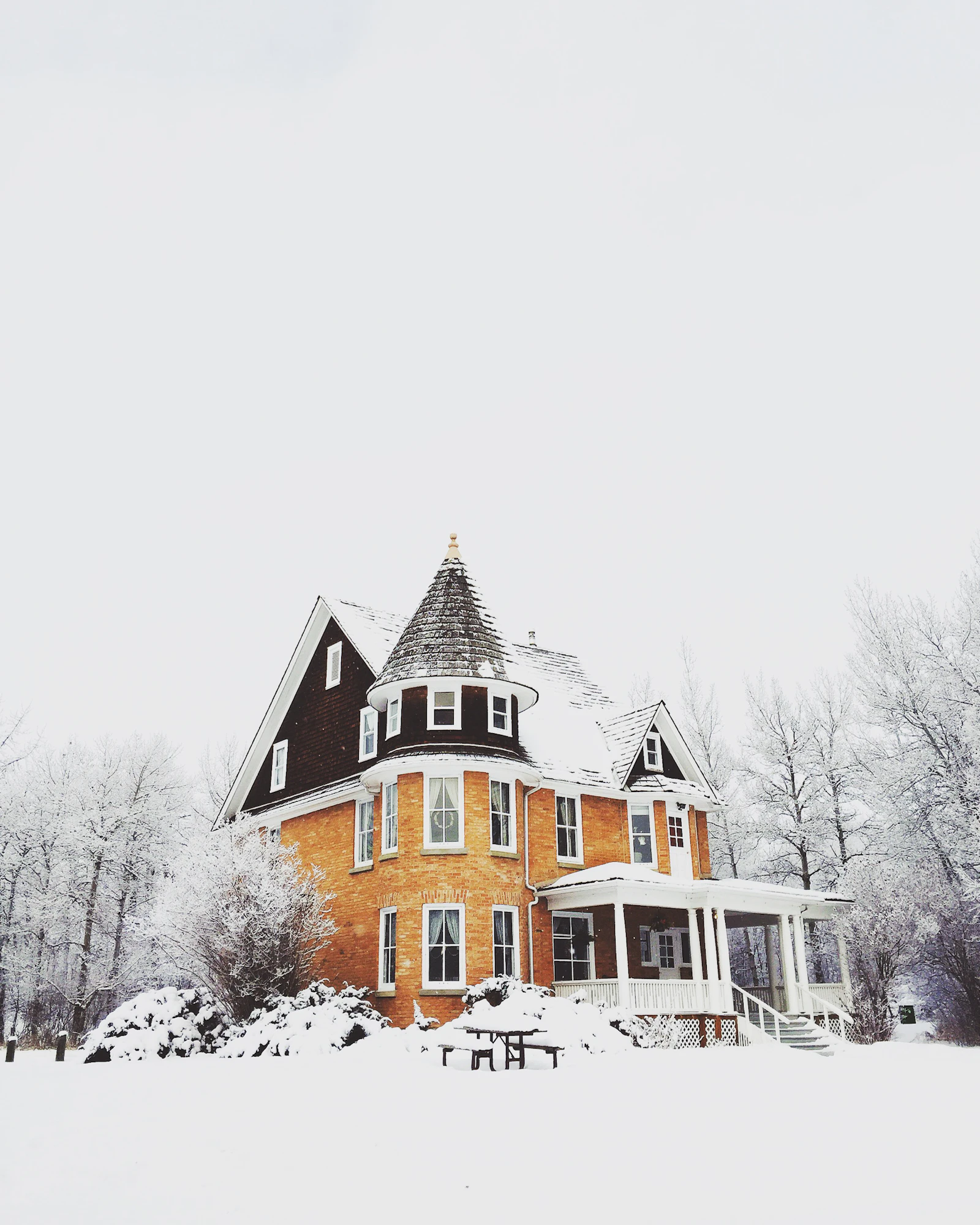 Victorian house with a shingled corner turret and wraparound porch surrounded by deep snow and frosted trees under a white sky