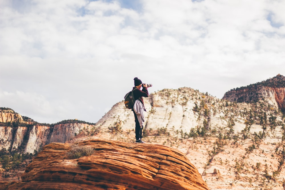 man wearing black jacket holding DSLR camera while taking photo standing on brown cliff during day time