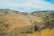 A series of rolling, dry hills covered with scattered vegetation and patches of trees stretches into the distance under a partly cloudy sky. The landscape is predominantly brown and green, with taller grasses visible in the foreground.