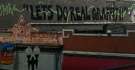 Graffiti saying 'LET'S DO REAL GRAFFITI' is painted on a building. The building has an ornate architectural design with a visible sign for a 'Foot and Ankle Clinic'. There are also street lamps visible in the foreground.