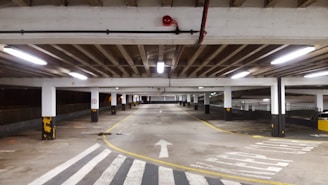 A large indoor parking garage with fluorescent lighting, concrete floors, and support columns is shown. The area appears empty, with a clear road for vehicles marked by arrows and a zebra crossing. Yellow and black safety markings are visible on the columns.