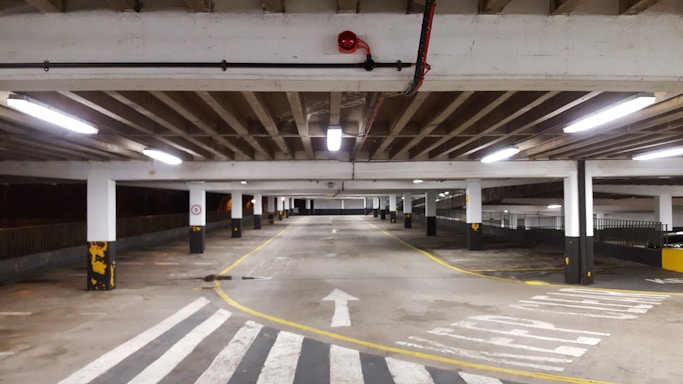A large indoor parking garage with fluorescent lighting, concrete floors, and support columns is shown. The area appears empty, with a clear road for vehicles marked by arrows and a zebra crossing. Yellow and black safety markings are visible on the columns.