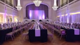 A grand ballroom set up for a formal event with elegant chandeliers hanging from a high ceiling. Long tables are neatly arranged with white tablecloths and centered with decorative branches. The room is softly lit with ambient purple lighting, creating a sophisticated atmosphere. Chairs are placed uniformly around the tables, and a stage is visible at the far end.