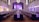 A grand ballroom set up for a formal event with elegant chandeliers hanging from a high ceiling. Long tables are neatly arranged with white tablecloths and centered with decorative branches. The room is softly lit with ambient purple lighting, creating a sophisticated atmosphere. Chairs are placed uniformly around the tables, and a stage is visible at the far end.