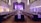 A grand ballroom set up for a formal event with elegant chandeliers hanging from a high ceiling. Long tables are neatly arranged with white tablecloths and centered with decorative branches. The room is softly lit with ambient purple lighting, creating a sophisticated atmosphere. Chairs are placed uniformly around the tables, and a stage is visible at the far end.