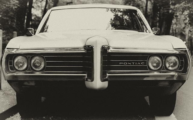 A vintage car with a prominent front grille and circular headlights, featuring the word 'Pontiac' in the center. The car is set against a natural, wooded background. The image has a sepia tone, giving it a nostalgic, classic feel.