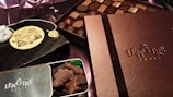 An assortment of luxury chocolates and treats are displayed. Boxes labeled Lenôtre Paris and Mariage Frères are prominently featured, suggesting high-end products. A selection of chocolates is arranged neatly on what appears to be a silk fabric background.
