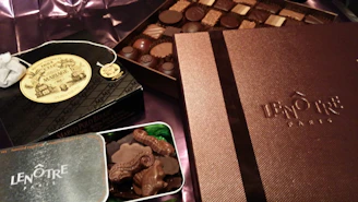 An assortment of luxury chocolates and treats are displayed. Boxes labeled Lenôtre Paris and Mariage Frères are prominently featured, suggesting high-end products. A selection of chocolates is arranged neatly on what appears to be a silk fabric background.