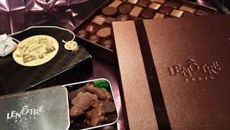 An assortment of luxury chocolates and treats are displayed. Boxes labeled Lenôtre Paris and Mariage Frères are prominently featured, suggesting high-end products. A selection of chocolates is arranged neatly on what appears to be a silk fabric background.