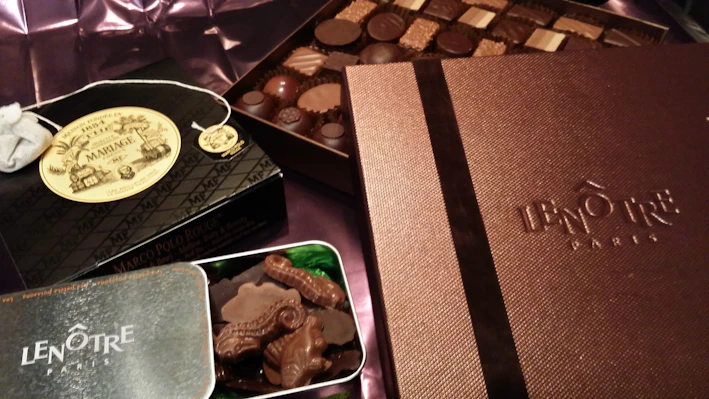 An assortment of luxury chocolates and treats are displayed. Boxes labeled Lenôtre Paris and Mariage Frères are prominently featured, suggesting high-end products. A selection of chocolates is arranged neatly on what appears to be a silk fabric background.