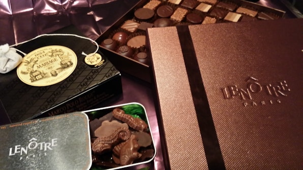 An assortment of luxury chocolates and treats are displayed. Boxes labeled Len&ocirc;tre Paris and Mariage Fr&egrave;res are prominently featured, suggesting high-end products. A selection of chocolates is arranged neatly on what appears to be a silk fabric background.