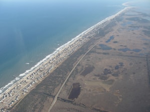 An aerial view showcasing a long, narrow strip of land bordered by the ocean on one side and marshy wetlands on the other. The land strip contains a densely packed row of small houses running parallel to the coastline. A road runs alongside the row of houses, cutting through the landscape.