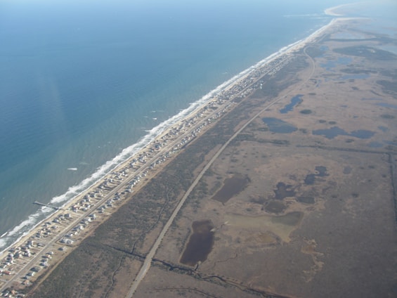 An aerial view showcasing a long, narrow strip of land bordered by the ocean on one side and marshy wetlands on the other. The land strip contains a densely packed row of small houses running parallel to the coastline. A road runs alongside the row of houses, cutting through the landscape.