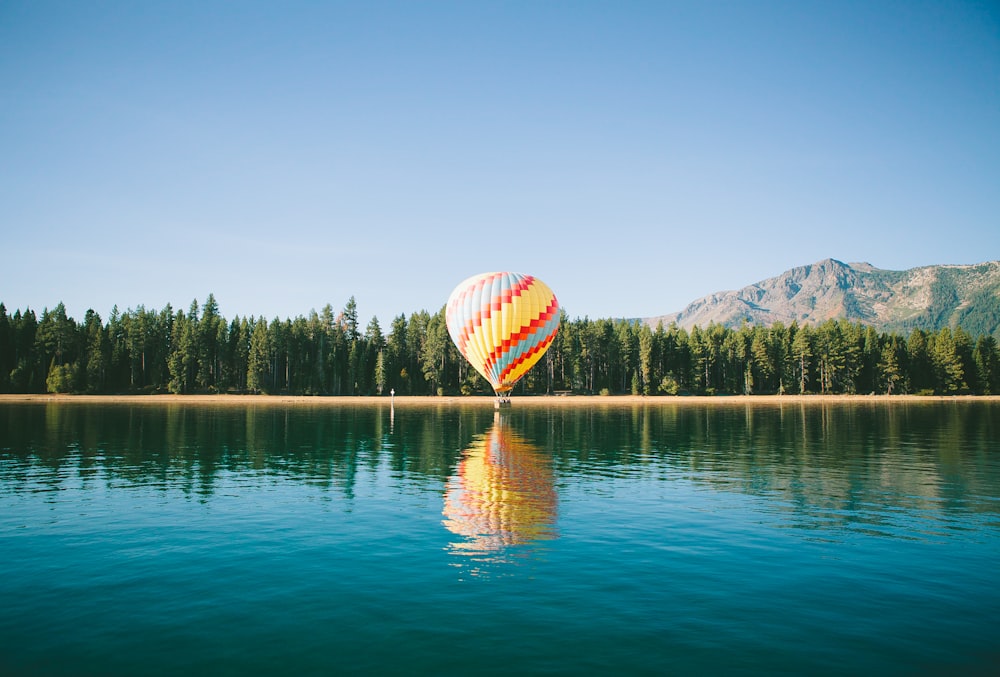 yellow, red, and blue hot air balloon near trees and body of water under blue sky during daytime