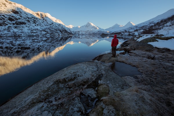 Man in red jacket looking over still lake with snowy mountains in background.