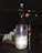 A translucent glass bottle prominently displays a label indicating it contains pure still water. Next to the bottle, a glass with a lit candle inside casts a warm light, creating a cozy ambiance. The background features subtle reflections and shadows, suggesting a dimly lit setting.