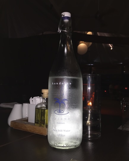 A translucent glass bottle prominently displays a label indicating it contains pure still water. Next to the bottle, a glass with a lit candle inside casts a warm light, creating a cozy ambiance. The background features subtle reflections and shadows, suggesting a dimly lit setting.