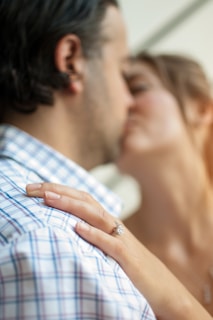 A romantic scene features a couple engaged in a close embrace, with a focus on a woman's hand resting on the man's shoulder. The woman's hand is adorned with a prominent diamond ring. The couple's faces are partially blurred, enhancing the intimate atmosphere.
