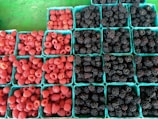 An array of berry containers filled with fresh raspberries and blackberries are neatly arranged in rows. The containers are light blue, contrasting with the vibrant red and dark purple berries. The background features a green surface, enhancing the colorful display of the fruit.