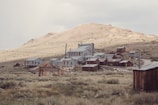 A collection of old, abandoned wooden and metal buildings sits in a desolate, grassy landscape beneath a barren hillside. The structures appear weathered and rustic, reminiscent of a ghost town from the past.