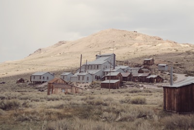 A collection of old, abandoned wooden and metal buildings sits in a desolate, grassy landscape beneath a barren hillside. The structures appear weathered and rustic, reminiscent of a ghost town from the past.