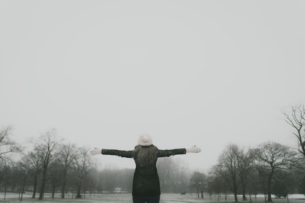 woman in black long-sleeved shirt standing and facing trees with stretched arms under white sky