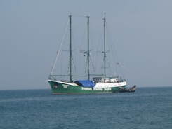 A large ship with three masts sails on calm waters. The ship is named 'Rongdhonu Friendship Hospital' and appears to be equipped for its purpose, suggesting a hospital ship. The sky is clear with a light blue tone, and there is a smaller boat docked alongside the main vessel.