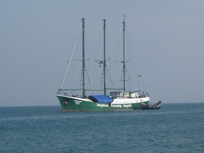 A large ship with three masts sails on calm waters. The ship is named 'Rongdhonu Friendship Hospital' and appears to be equipped for its purpose, suggesting a hospital ship. The sky is clear with a light blue tone, and there is a smaller boat docked alongside the main vessel.