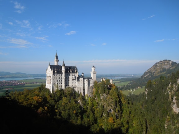 A fairytale-like castle stands majestically atop a lush, forested hill, surrounded by a picturesque landscape of green fields and rolling mountains under a clear blue sky.