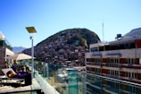 A hillside densely packed with colorful houses is visible in the background, suggesting a favela community. In the foreground, a rooftop with glass railings has lounge chairs, where people are relaxing under the sun beside a swimming pool. A solar panel and a large umbrella provide some shade.