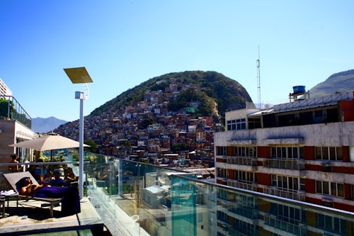 A hillside densely packed with colorful houses is visible in the background, suggesting a favela community. In the foreground, a rooftop with glass railings has lounge chairs, where people are relaxing under the sun beside a swimming pool. A solar panel and a large umbrella provide some shade.