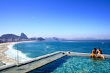 A couple sitting on the edge of a swimming pool overlooking a crowded beach and a deep blue sea