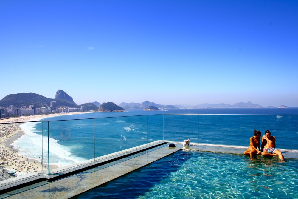 Um casal sentado à beira de uma piscina com vista para uma praia lotada e um mar azul profundo