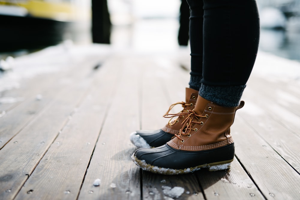 pessoa vestindo botas de pato de couro marrom e preto em pé na doca de madeira marrom