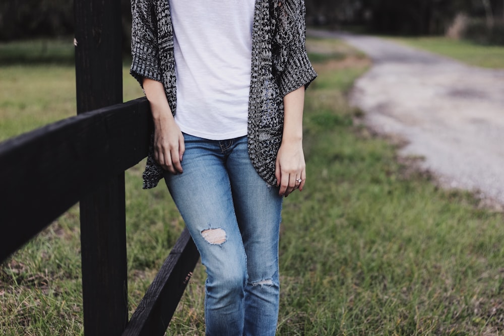 person wearing blue denim jeans and black cardigan leaning on black wooden fence beside road during daytime