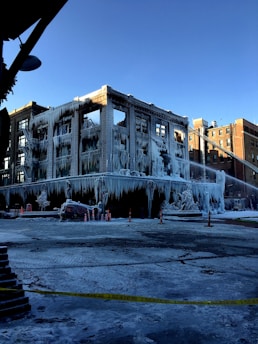 A partially burnt building covered in a thick layer of ice, with icicles hanging from various surfaces. Emergency cones are placed around the perimeter and a fire hose can be seen spraying water onto the structure. The ice gives the building a stark, frozen appearance. Yellow police tape cordons off the area, suggesting a recent fire incident.