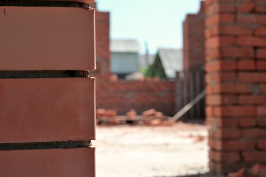 Close-up of a brick wall section with a background of a construction site, showing more brickwork in progress. The image is focused on the bricks with blurred background.