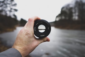 A hand holds a camera lens with a body of water and trees in the background. The view through the lens is focused on the distant landscape, while the surroundings appear blurred. The image suggests a perspective or focus theme.