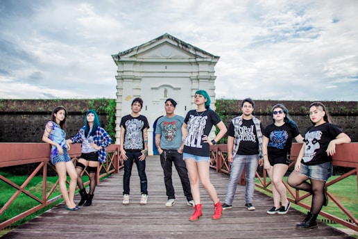 A group of young adults, casually dressed, poses confidently on a wooden walkway in front of a historic-looking building. The sky is overcast, adding drama to the scene. Each person showcases a unique style, with graphic T-shirts, varied hairstyles, and accessories.