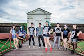A group of young adults, casually dressed, poses confidently on a wooden walkway in front of a historic-looking building. The sky is overcast, adding drama to the scene. Each person showcases a unique style, with graphic T-shirts, varied hairstyles, and accessories.
