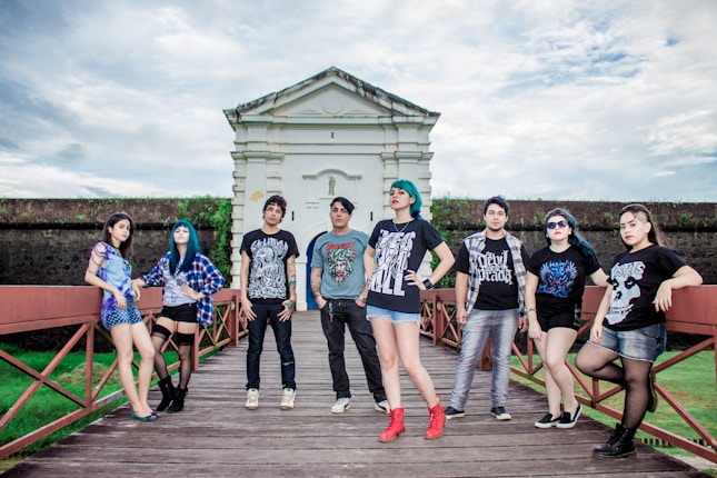 A group of young adults, casually dressed, poses confidently on a wooden walkway in front of a historic-looking building. The sky is overcast, adding drama to the scene. Each person showcases a unique style, with graphic T-shirts, varied hairstyles, and accessories.