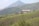A large building with a tin roof is situated in the midst of lush green tea plantations that cover rolling hills. In the background, a tall mountain rises, adding depth to the landscape.