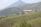 A large building with a tin roof is situated in the midst of lush green tea plantations that cover rolling hills. In the background, a tall mountain rises, adding depth to the landscape.