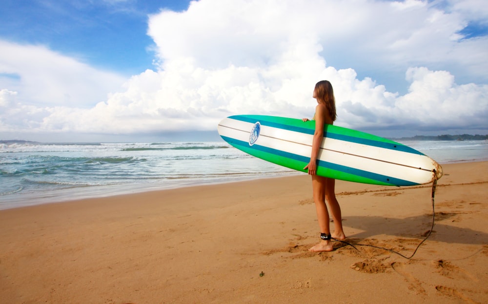 femme debout près de la mer tenant une planche de surf blanche, bleue et verte sous le ciel bleu