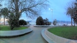 A peaceful waterfront park with a curved pathway, lined with trees and shrubs. Benches are integrated into the path design. A view of boats docked at a marina is visible, with mountains in the distance under a clear, evening sky. Street lamps are lit, suggesting dusk or dawn.